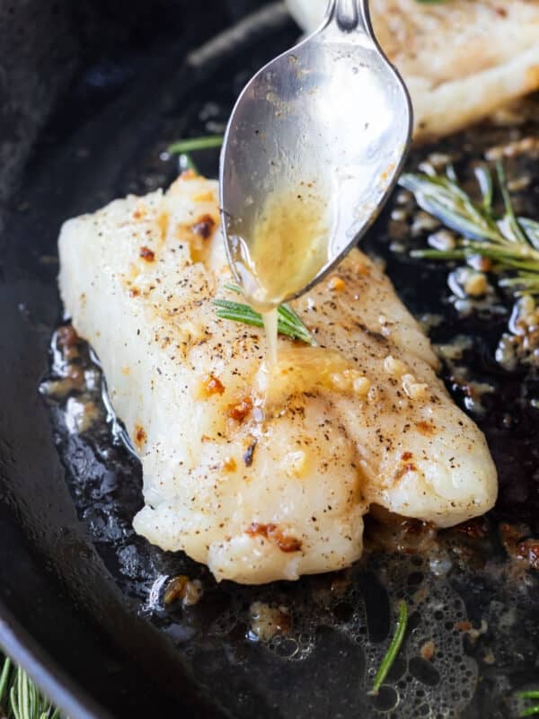 Garlic butter sauce is drizzled over a cod fillet in a cast iron skillet.