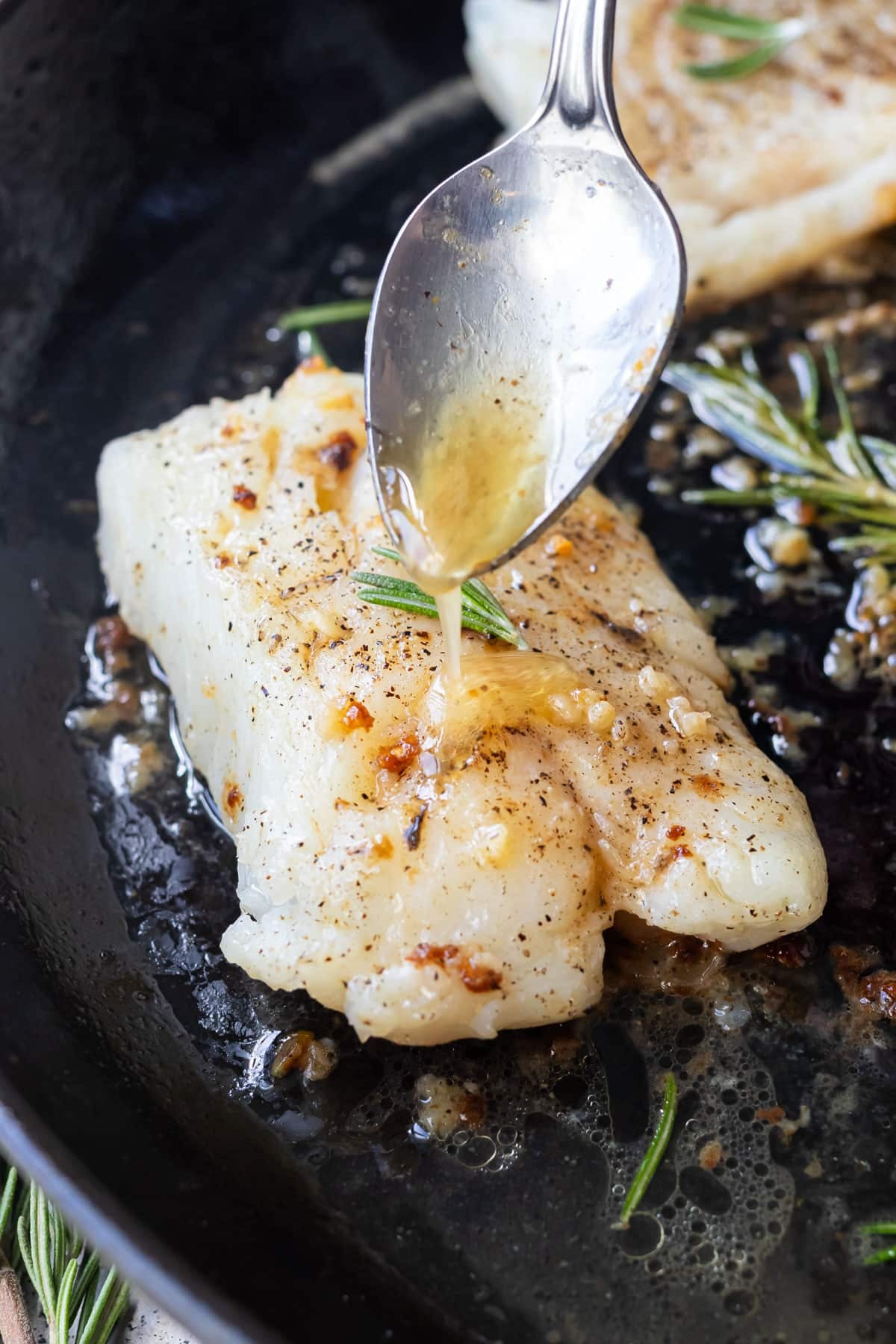 Spooning garlic rosemary butter over cod fillets in a skillet.