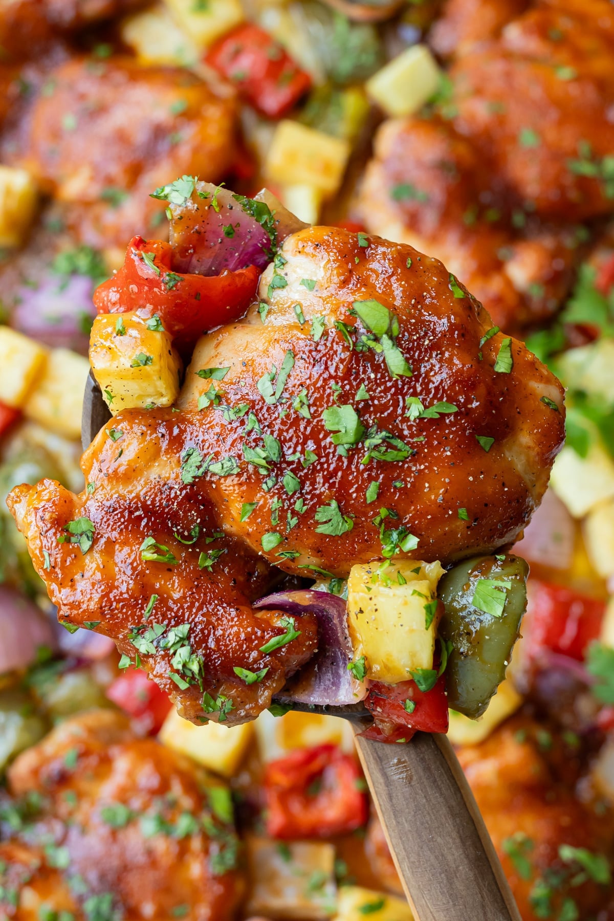 A spatula lifts up a caramelized Hawaiian Chicken thigh from a sheet pan with roasted pineapple and bell peppers.