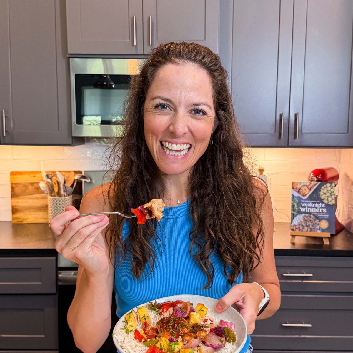 London Lea is in her Austin, Texas kitchen holding a bowl of sheet pan Hawaiian chicken and a bite of chicken and pineapple on a fork.