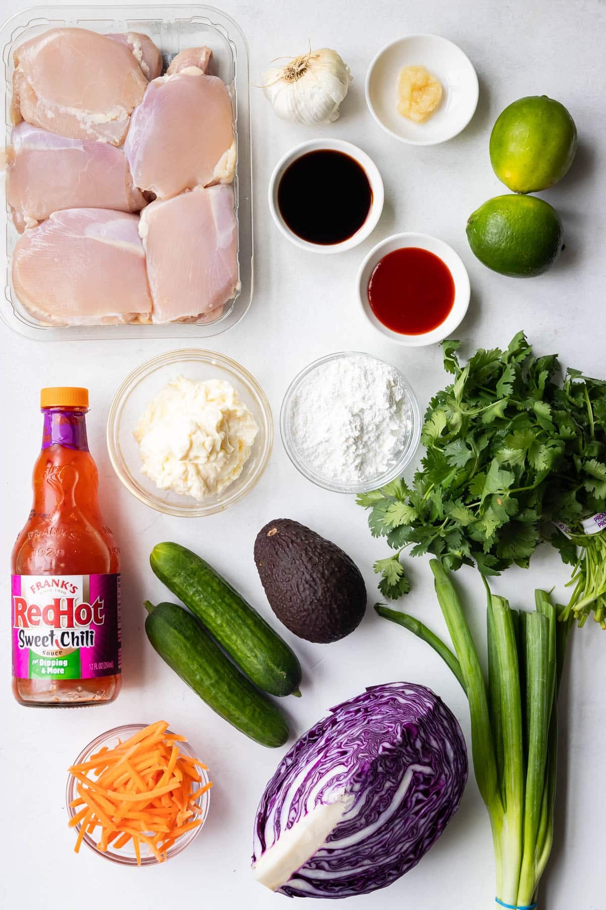 Ingredients for crispy bang bang chicken bowls laid out on a white counter, including chicken thighs, sweet chili sauce, cornstarch, and fresh vegetables.