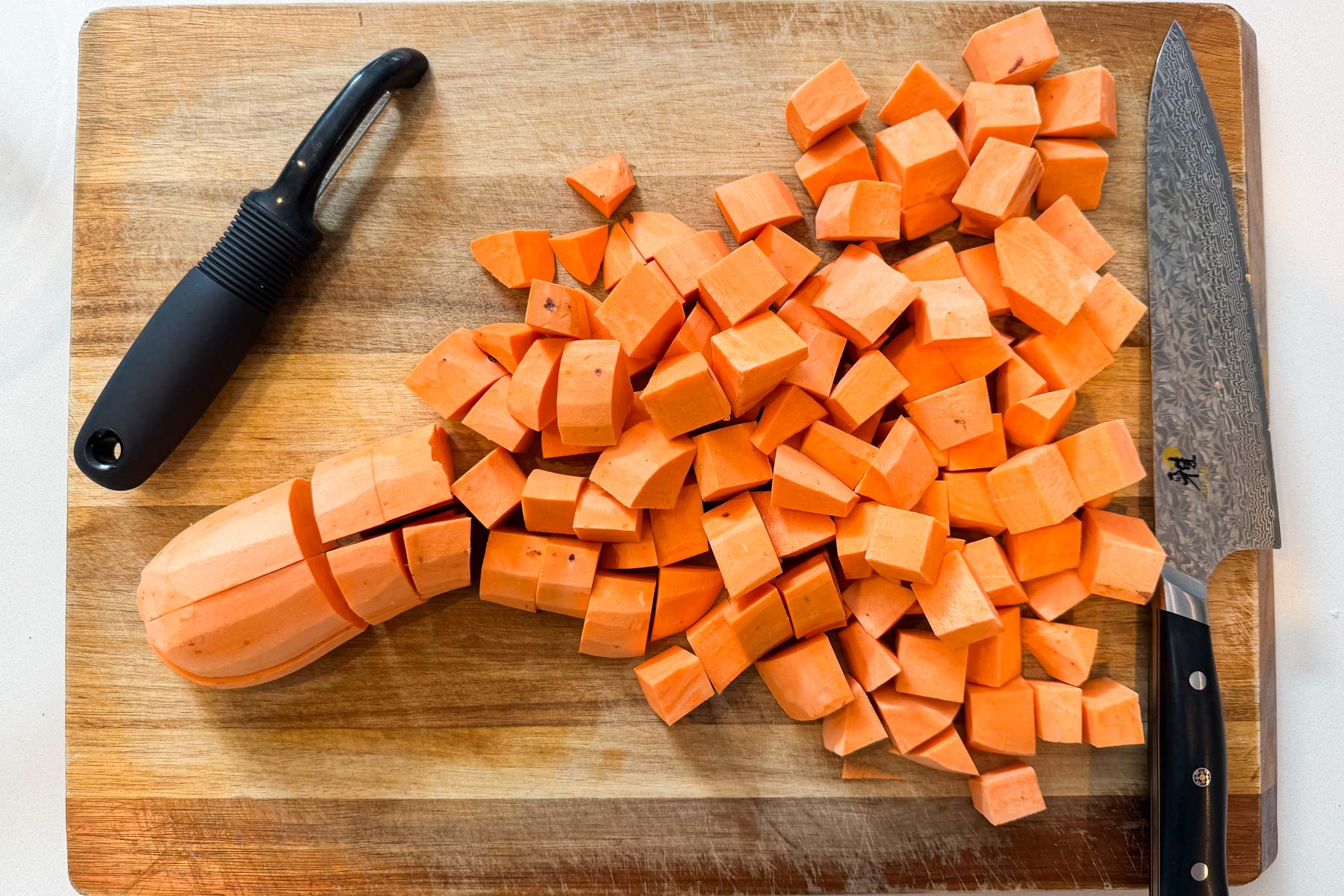 A sweet potato is peeled and cut into three-quarter-inch cubes on a wooden cutting board.