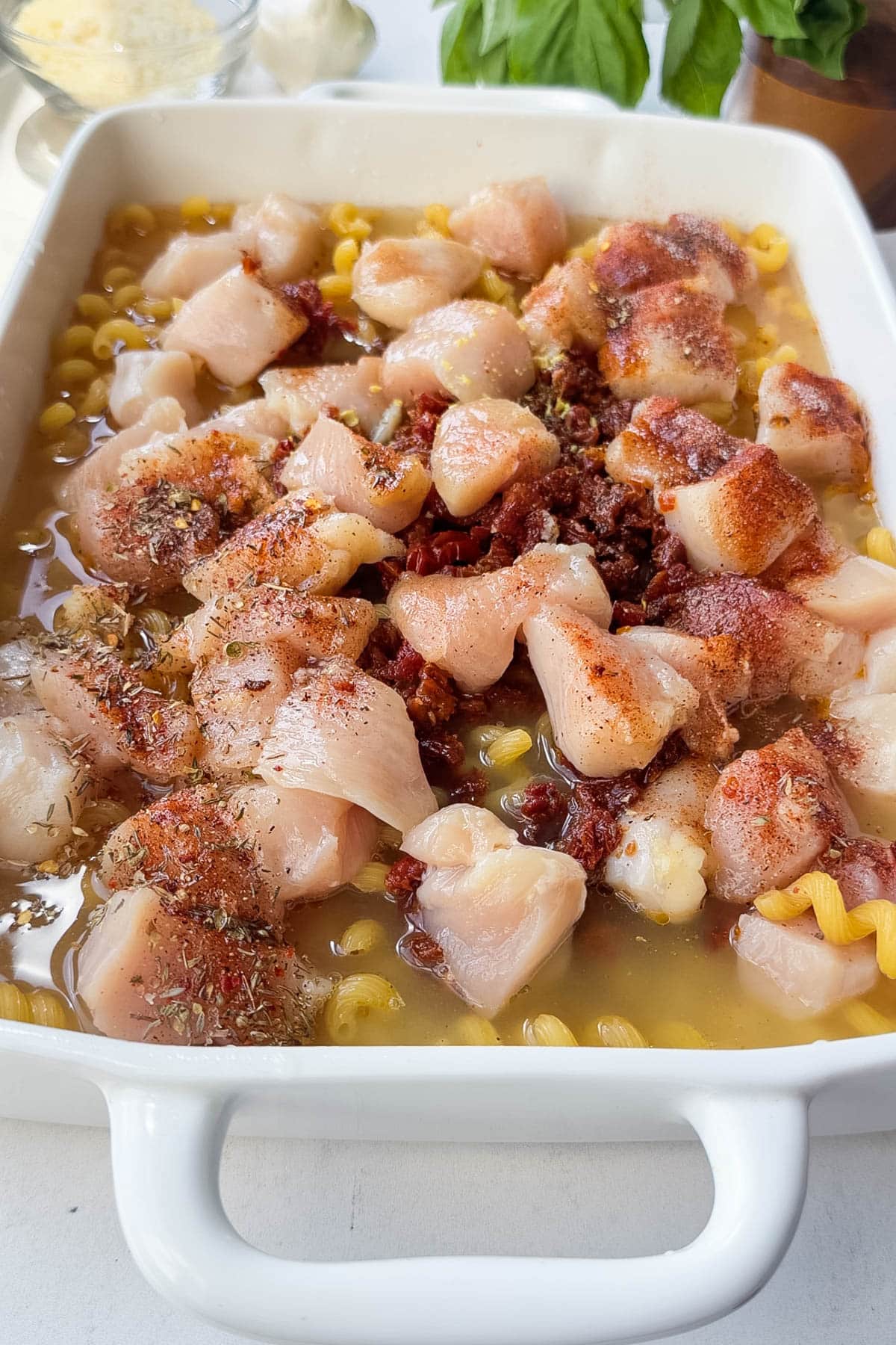 The chicken, seasonings, pasta, and broth are in a baking dish for a marry me chicken pasta dump and bake recipe.