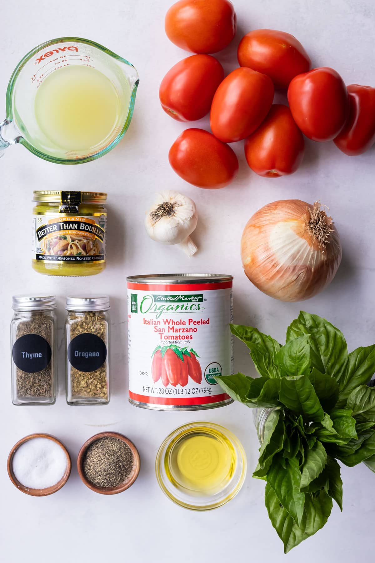 Roma tomatoes, a head of garlic, yellow onion, canned San Marzano tomatoes, fresh basil, olive oil, and seasonings arranged on a white counter