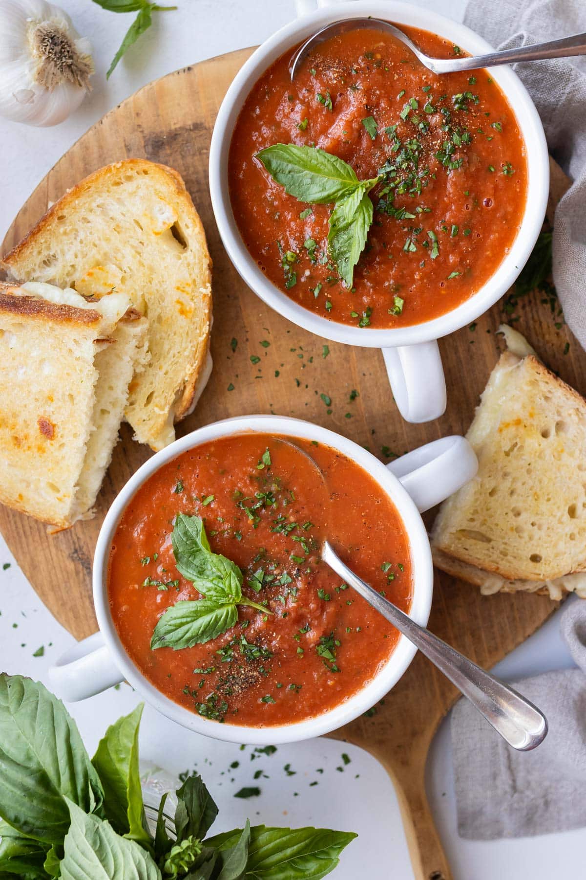 Two cups full of roasted tomato basil soup on a wooden board with grilled cheese sandwiches for dipping