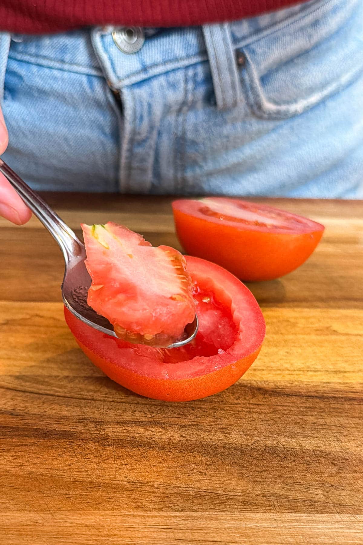 A spoon scooping the seeds and core out of a halved roma tomato on a wooden cutting board