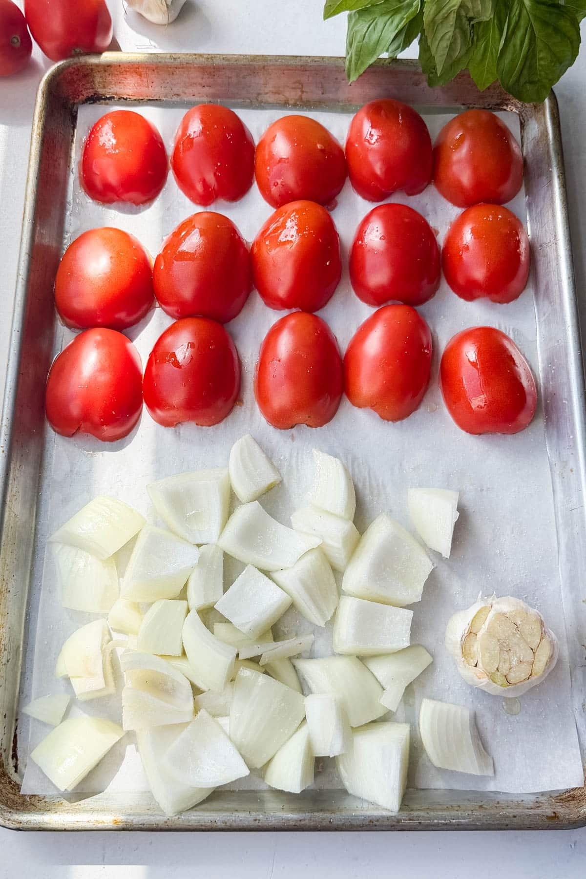 Halved roma tomatoes, chopped onion, and a trimmed head of garlic on a parchment-lined sheet pan before roasting