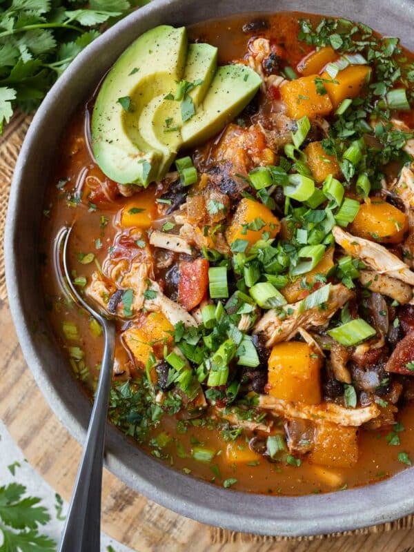 Bowl of chipotle chicken chili with butternut squash, avocado, and green onions on a wooden board