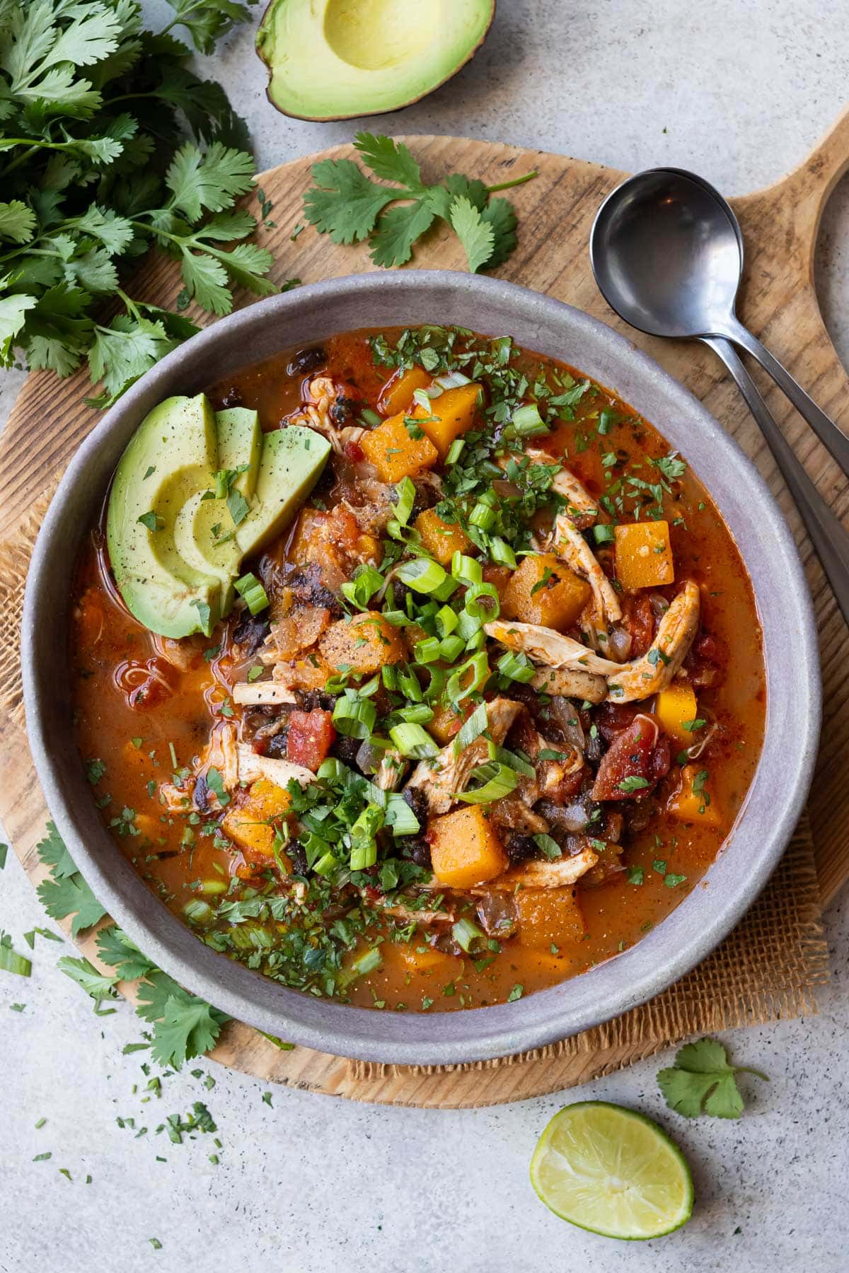 A bowl of chunky butternut squash chicken chili is served with avocado, green onions and cilantro.