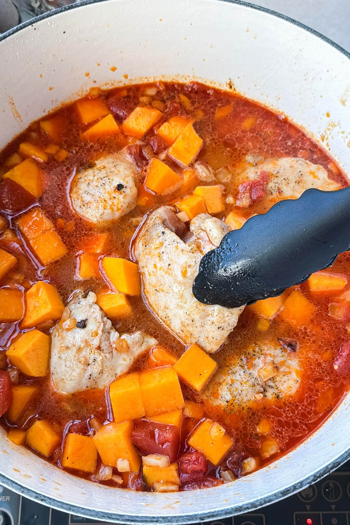 Tongs lifting chicken thighs out of simmering butternut squash chicken chili