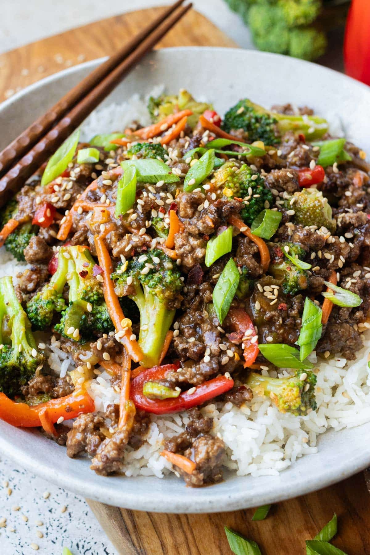 Ground beef and broccoli with bell peppers and carrots is served over rice with chopsticks in a white bowl.
