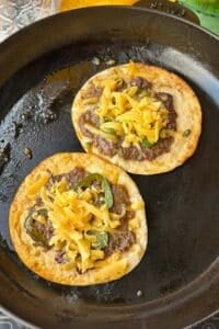 Flipped smashed tacos topped with shredded cheddar melting in the skillet