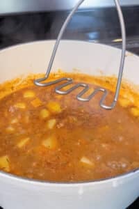 A potato masher lightly mashes potatoes in the pot to thicken shepherd's pie soup.