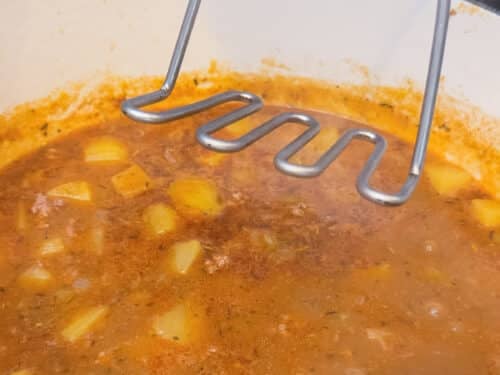 A potato masher lightly mashes potatoes in the pot to thicken shepherd's pie soup.